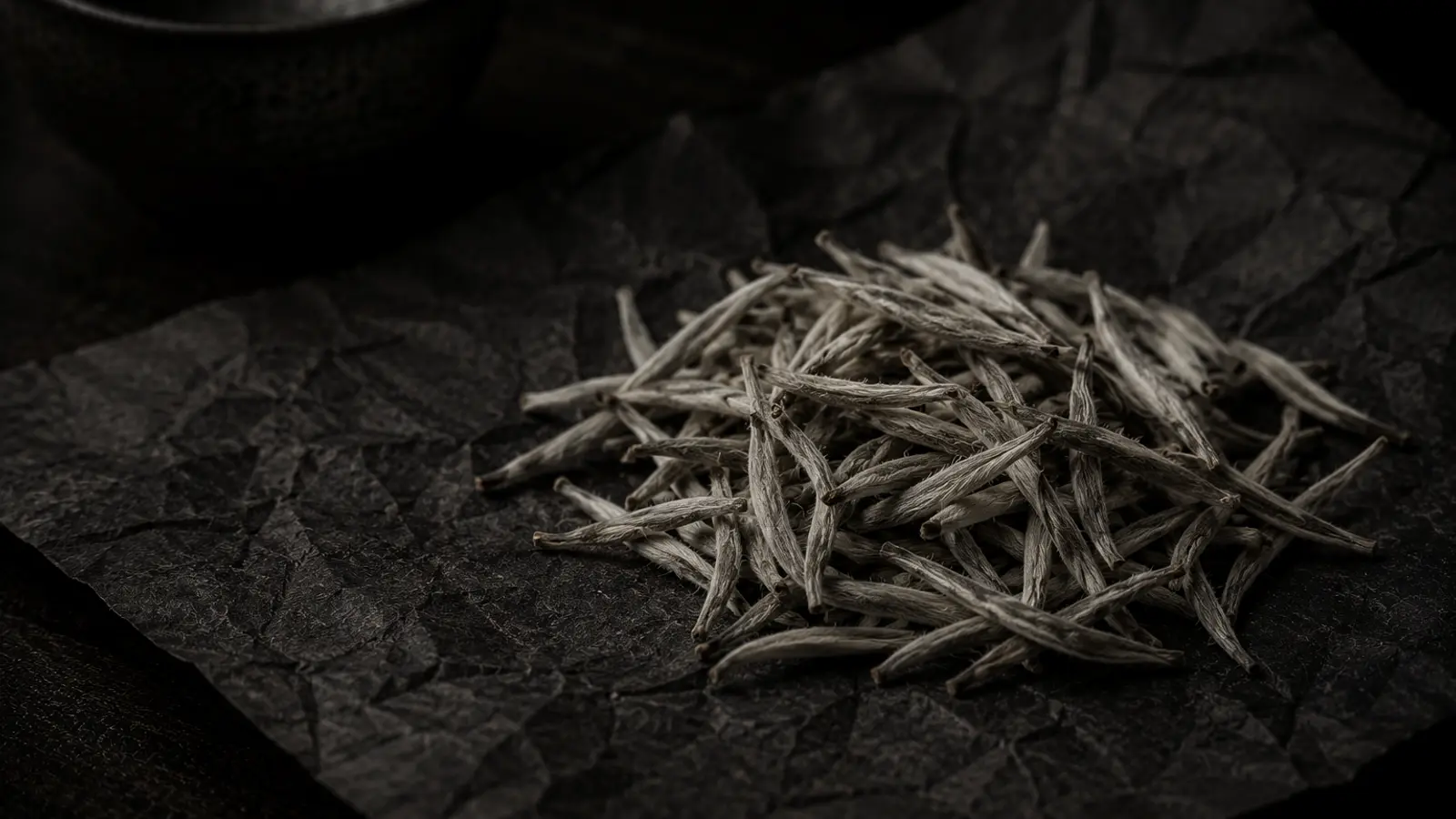 Silver needle white tea buds with fine white hairs beside a glass cup of pale golden liquor on marble, in soft natural light.